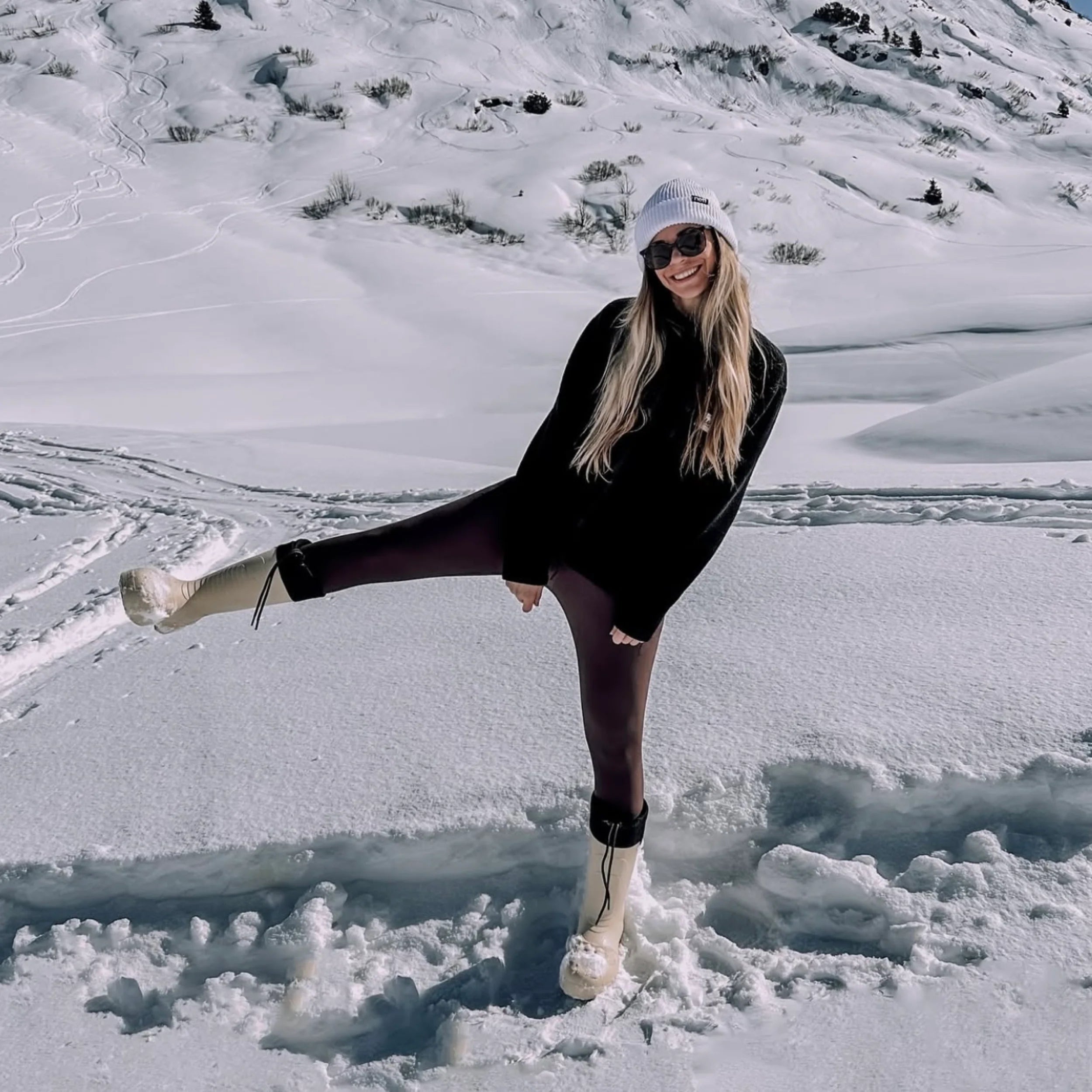 Person posing in the snow wearing a black outfit and white boots